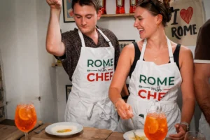 Two guests wearing Rome With Chef aprons laughing while making pasta alongside Aperol Spritz cocktails during a Carpe Diem Tours Rome spritz and spaghetti cooking class