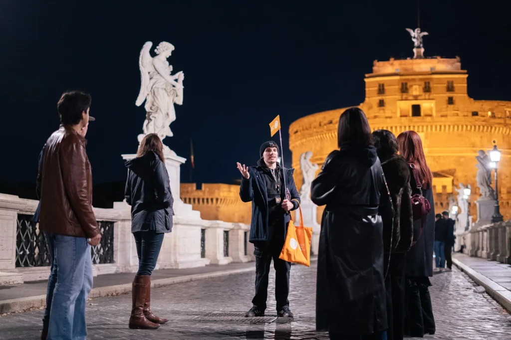 A tour guide on the Ponte Sant'Angelo with the illuminated Castel Sant'Angelo in the background during a night tour.